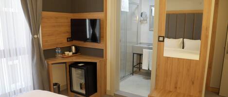 A hotel room with a bed, a TV, a minibar, a shower area, and a wooden wardrobe.