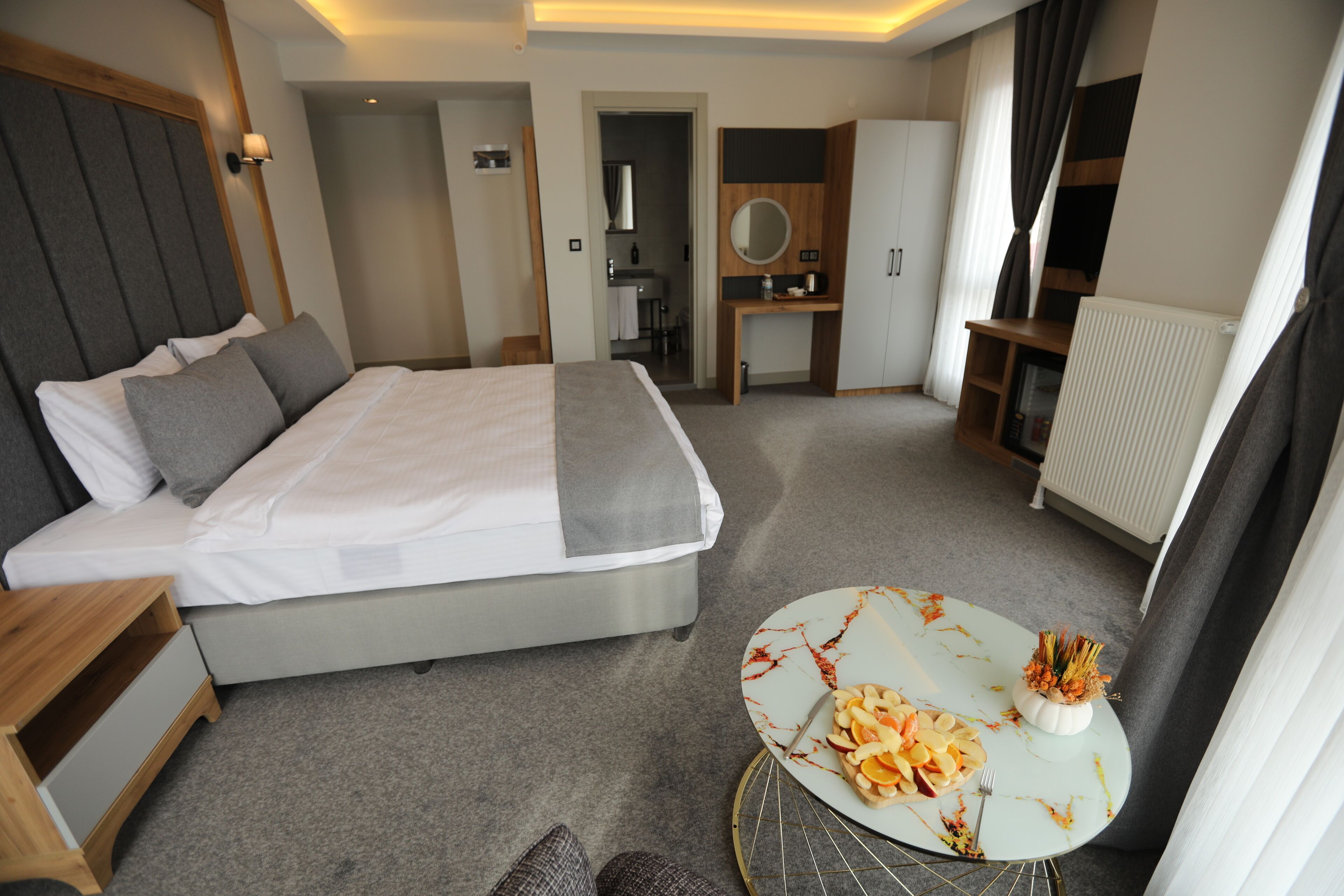 A modern hotel room with a large bed, a small desk, a round table with a plate of food, and a wardrobe.
