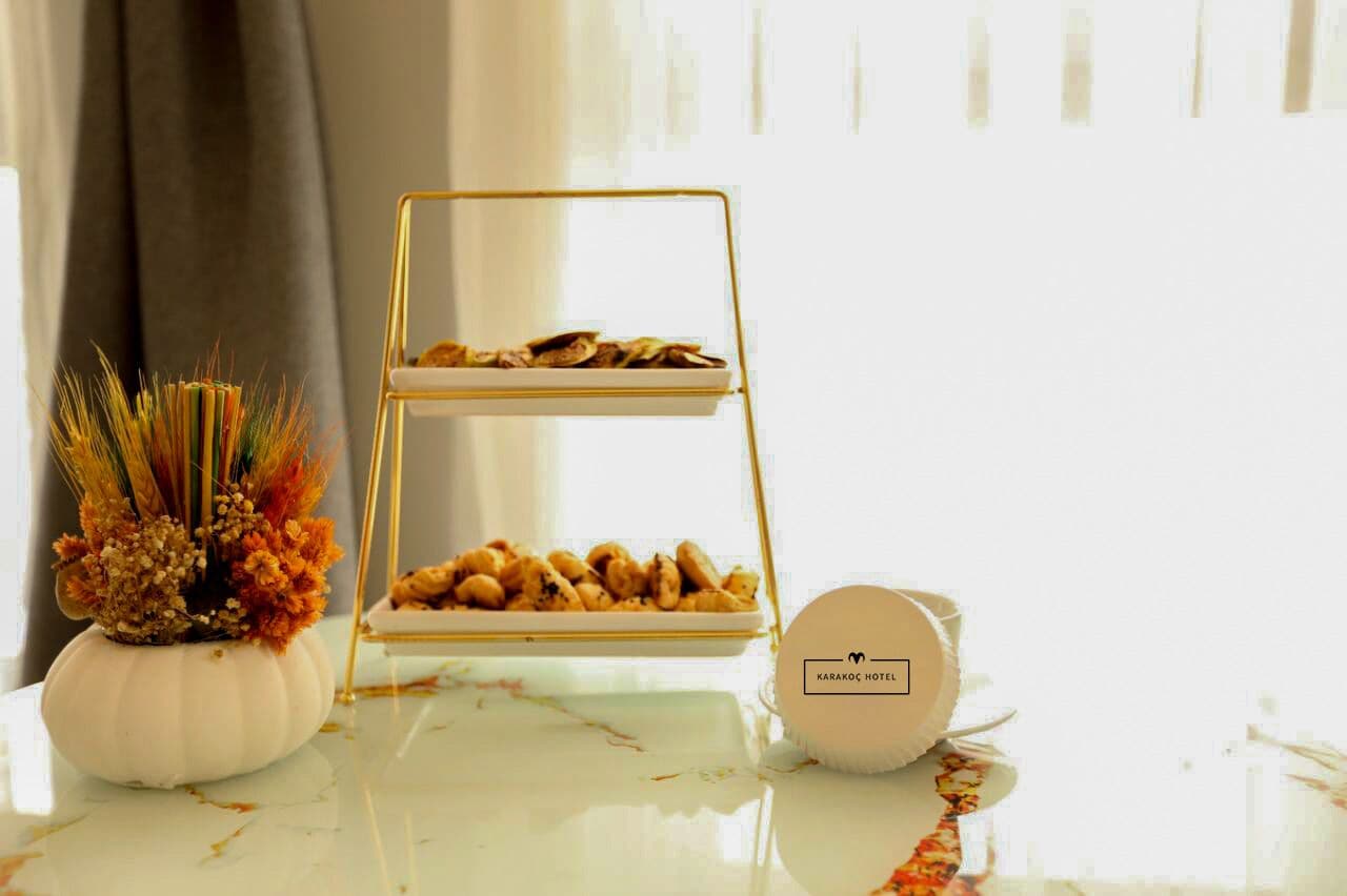 Un présentoir à deux niveaux, avec un cadre doré, sur lequel sont disposées des pâtisseries, est placé sur une table en marbre blanc. Il est accompagné d’un vase blanc contenant des fleurs séchées et d’un récipient rond portant le nom de l’hôtel.