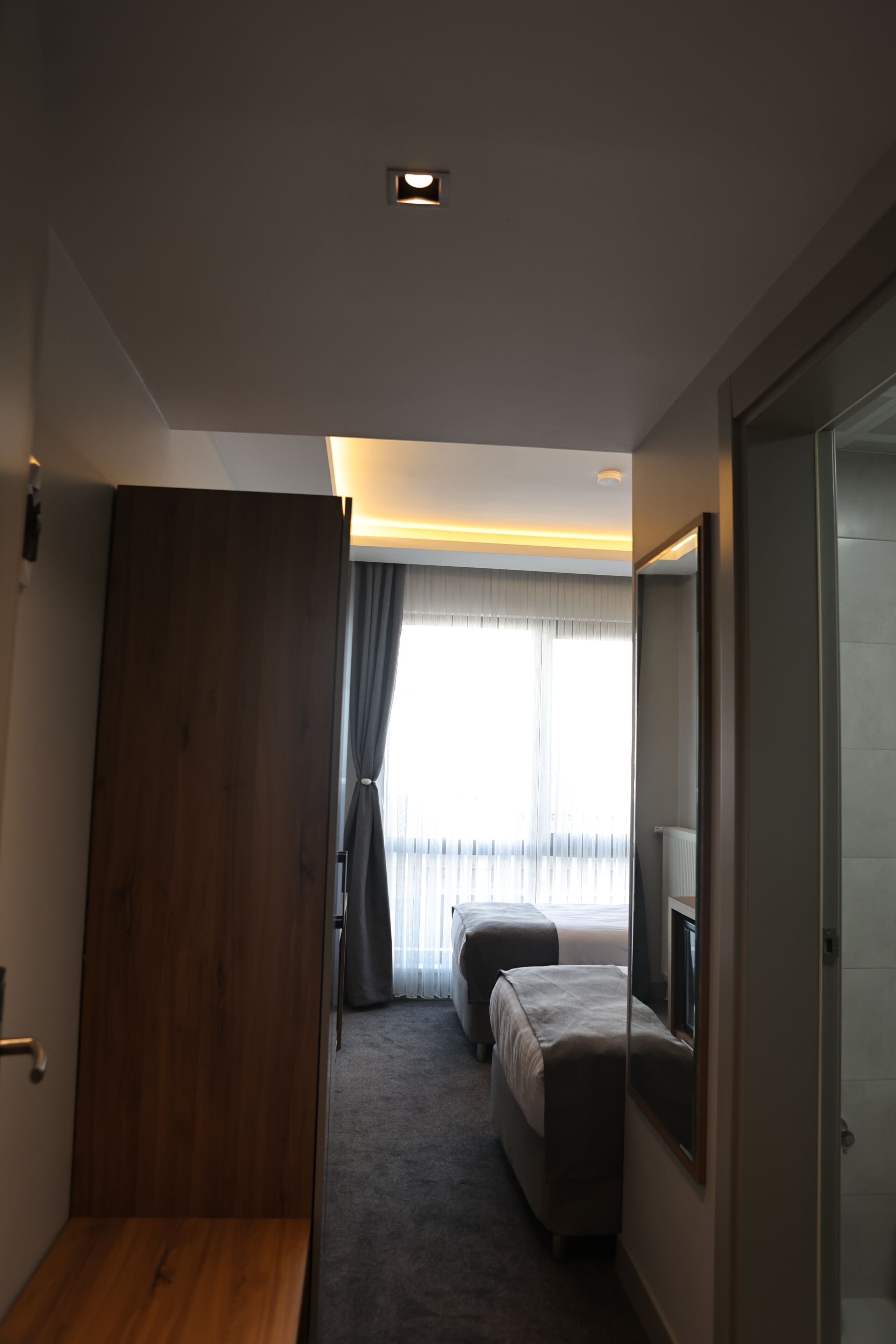 A modern hotel room with a wooden wardrobe, a bed with a grey headboard, a large mirror, and a window with sheer curtains.