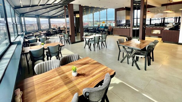 A modern dining area with multiple tables and chairs, large windows offering a city view, and a high ceiling with recessed lighting. - KARAKOÇ HOTEL  (Van)