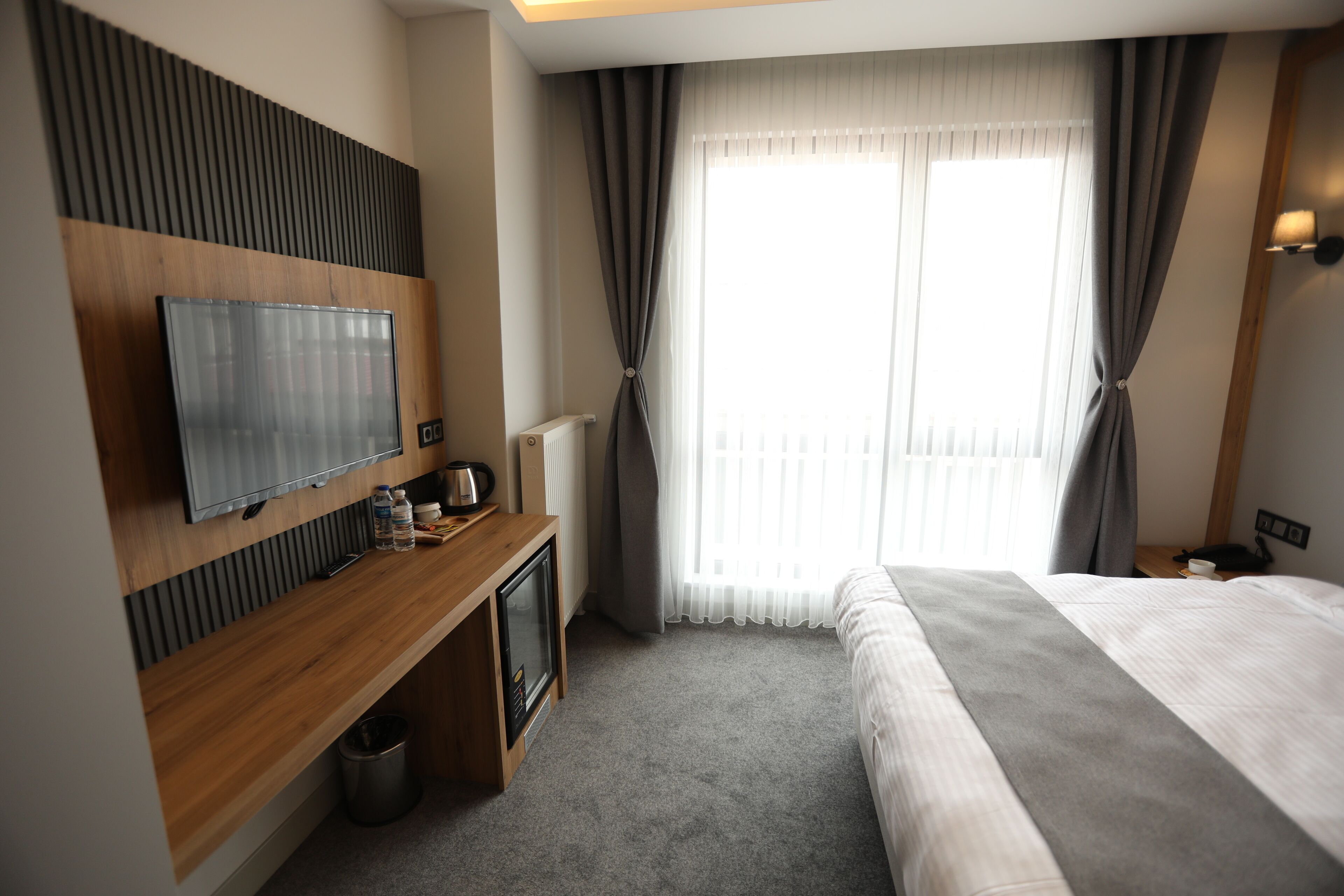 Una habitación de hotel con una cama, un televisor montado en un panel de madera, un escritorio pequeño con un hervidor y una ventana con cortinas.