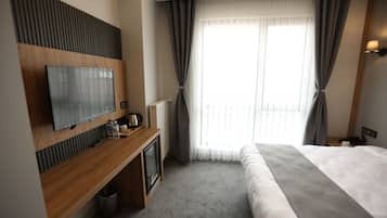 A hotel room with a bed, a TV mounted on a wooden panel, a small desk with a kettle, and a window with curtains.