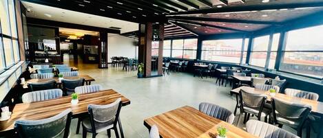 Un restaurante moderno con mesas y sillas de madera, grandes ventanales que ofrecen vistas a la ciudad y un alto techo con iluminación lineal.