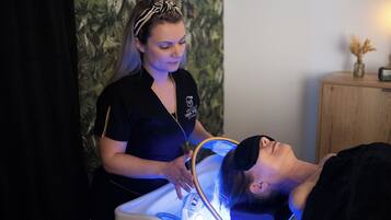 A person lying in a treatment chair with a headlamp on, a therapist standing beside them, and a wooden cabinet with a vase and clock in the background.