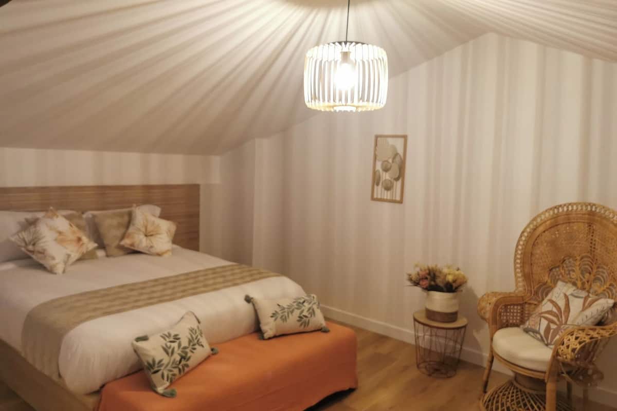 A bedroom with a large bed, a wooden headboard, an orange bench, a wicker chair, a hanging light fixture, and a small table with a vase.