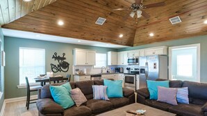 A spacious living room with a high ceiling, wooden beams, and a ceiling fan. It includes a brown leather sofa set, a wooden coffee table, and a dining area with a table and chairs. - Angler’s Paradise! Amazing fishing w/ in minutes! 3 bed,sleeps 7 ppl, 2 bath. (Hackberry)