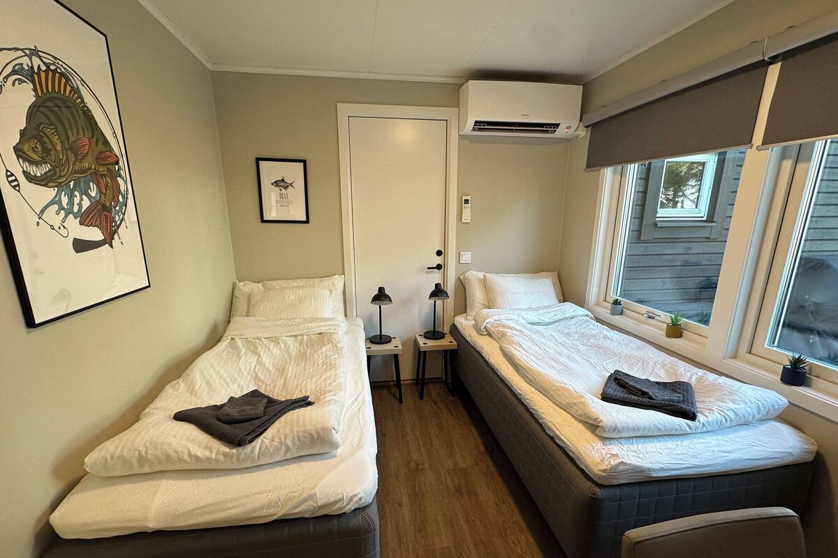 A compact room with two single beds, a small bedside table with a lamp, a framed picture on the wall, and a window with a view of the outside.