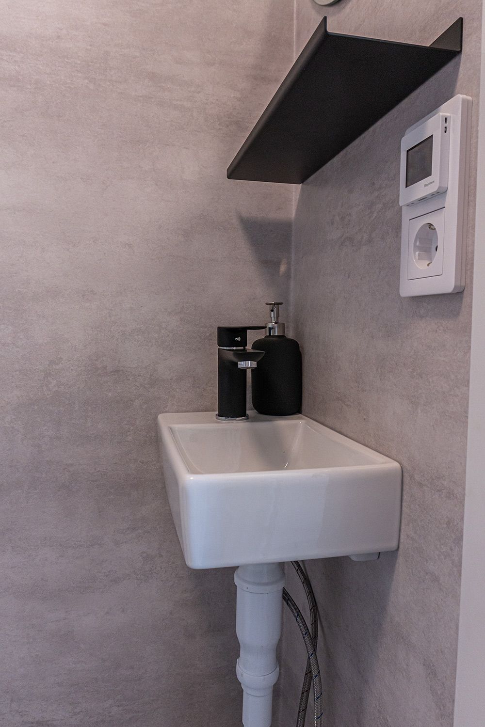 Un baño con un lavamanos blanco, un dispensador de jabón negro y un pequeño dispositivo blanco en la pared.