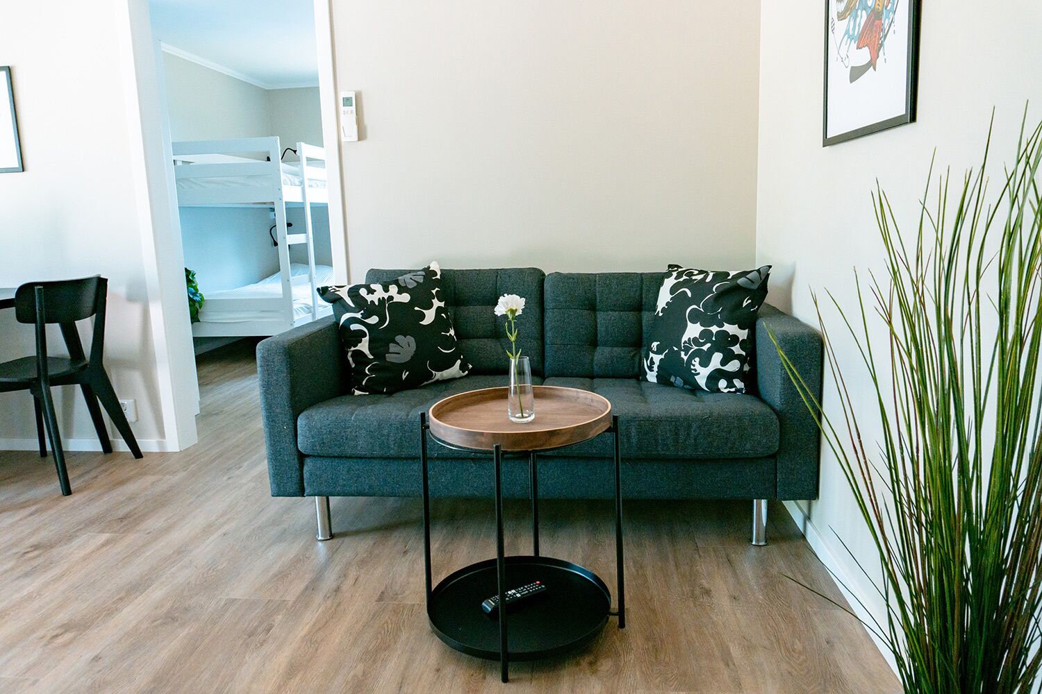 A modern living room with a grey sofa, two patterned cushions, a small wooden side table with a vase, and a remote control on a circular stand.