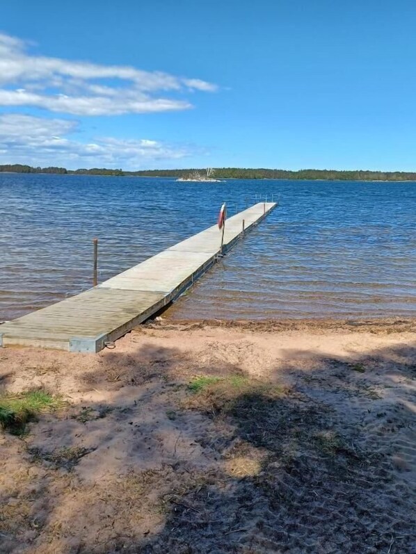 A wooden pier extending into a calm lake with a flag at the end, surrounded by a sandy shore and trees. - Perchlodge (Monsteras)