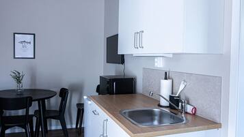 A compact kitchen with a wooden countertop, a stainless steel sink, and white cabinets. There is a small dining table with chairs and a framed picture on the wall.