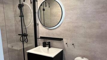A modern bathroom with a glass-enclosed shower, a white sink, a toilet, and a round mirror.