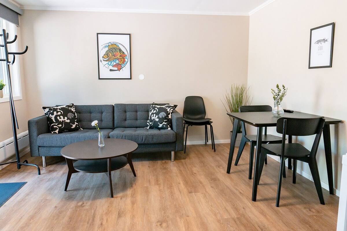 A modern living room with a grey sofa, a round coffee table, and a dining table with chairs. There are two framed pictures on the walls.