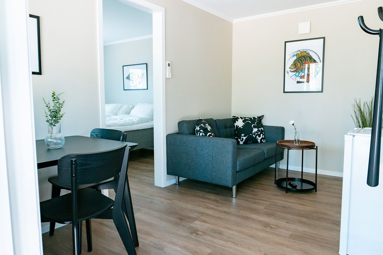 A modern living room with a grey sofa, a dining table with chairs, a small round table with a vase, and a bed in the background.