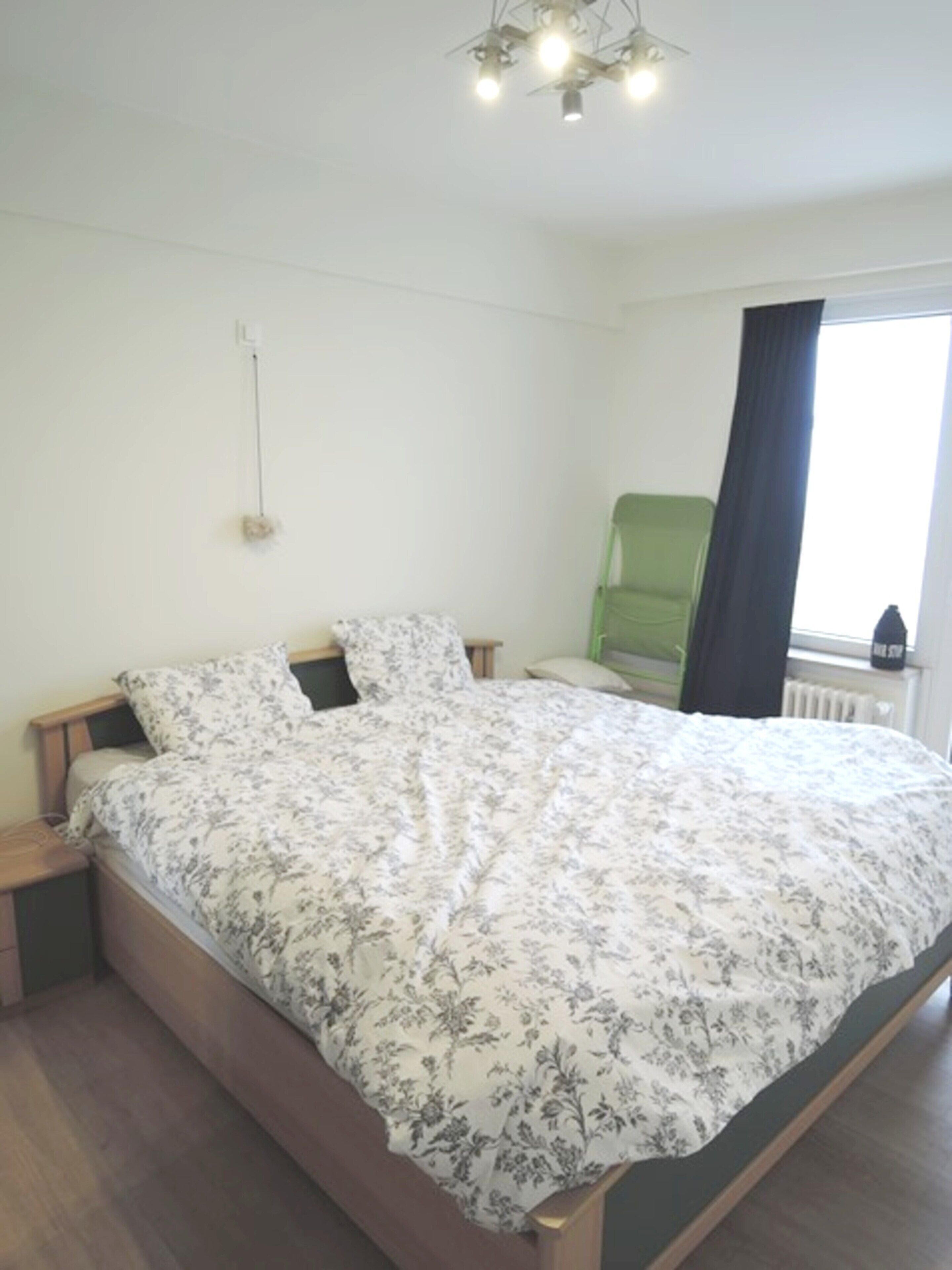 A bedroom with a wooden bed, floral bedding, a green chair, and a window with curtains.