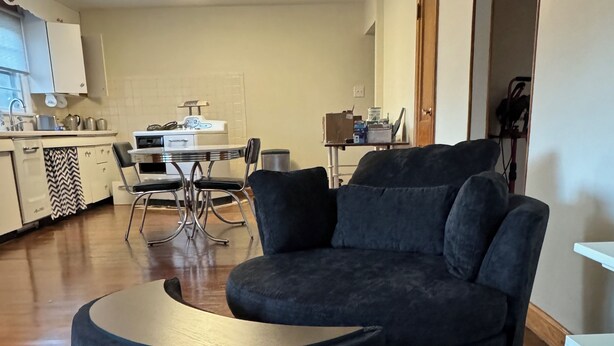 Un salon avec un canapé d’angle bleu foncé, un pouf rond, une table à manger avec des chaises, et un coin cuisine à l’arrière-plan.