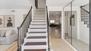 Una escalera con acabado de madera, paredes blancas y diseño moderno. Se observa un sofá y una zona de comedor al fondo.