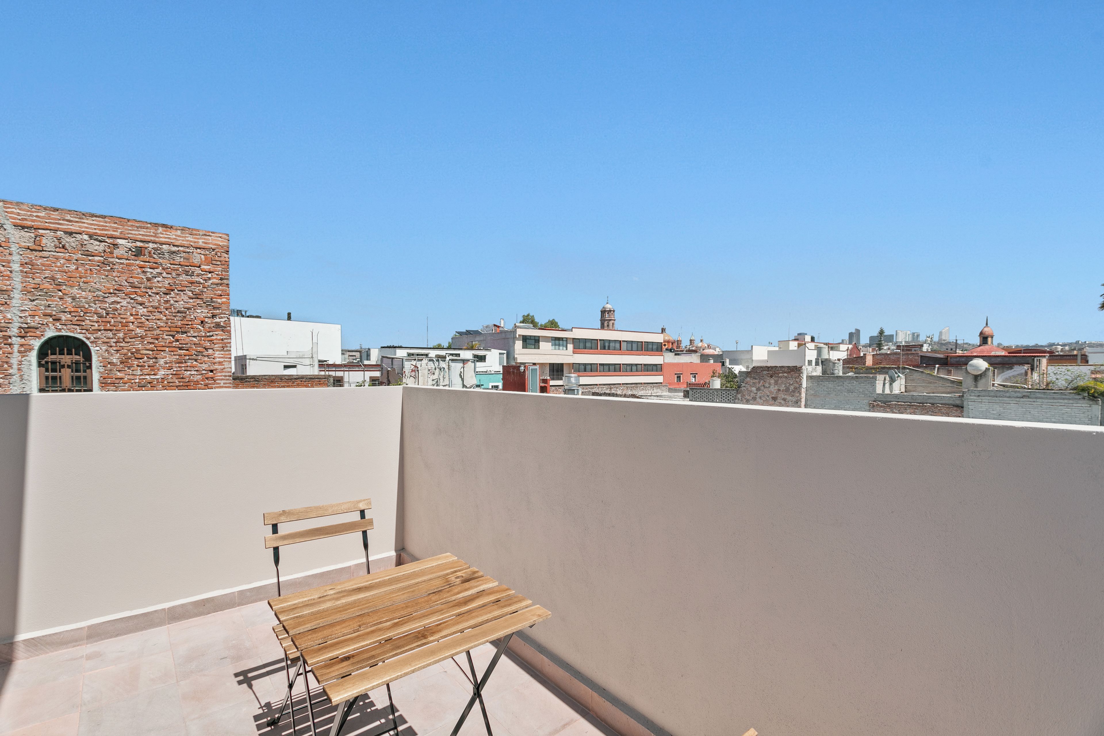 A rooftop terrace with a wooden bench, a view of the cityscape, and a brick building.