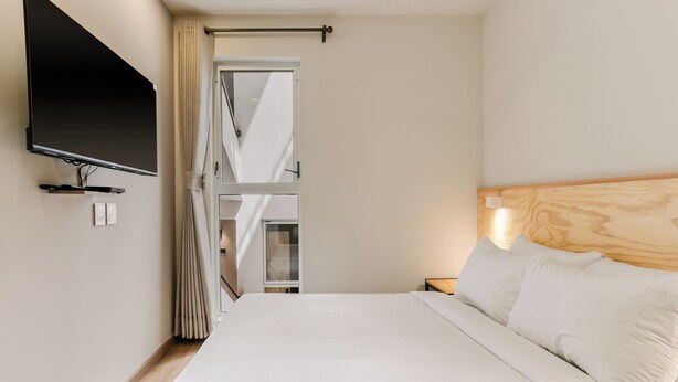 Habitación de hotel con cama, televisión montada en la pared, cabecera de madera y ventana con cortinas.