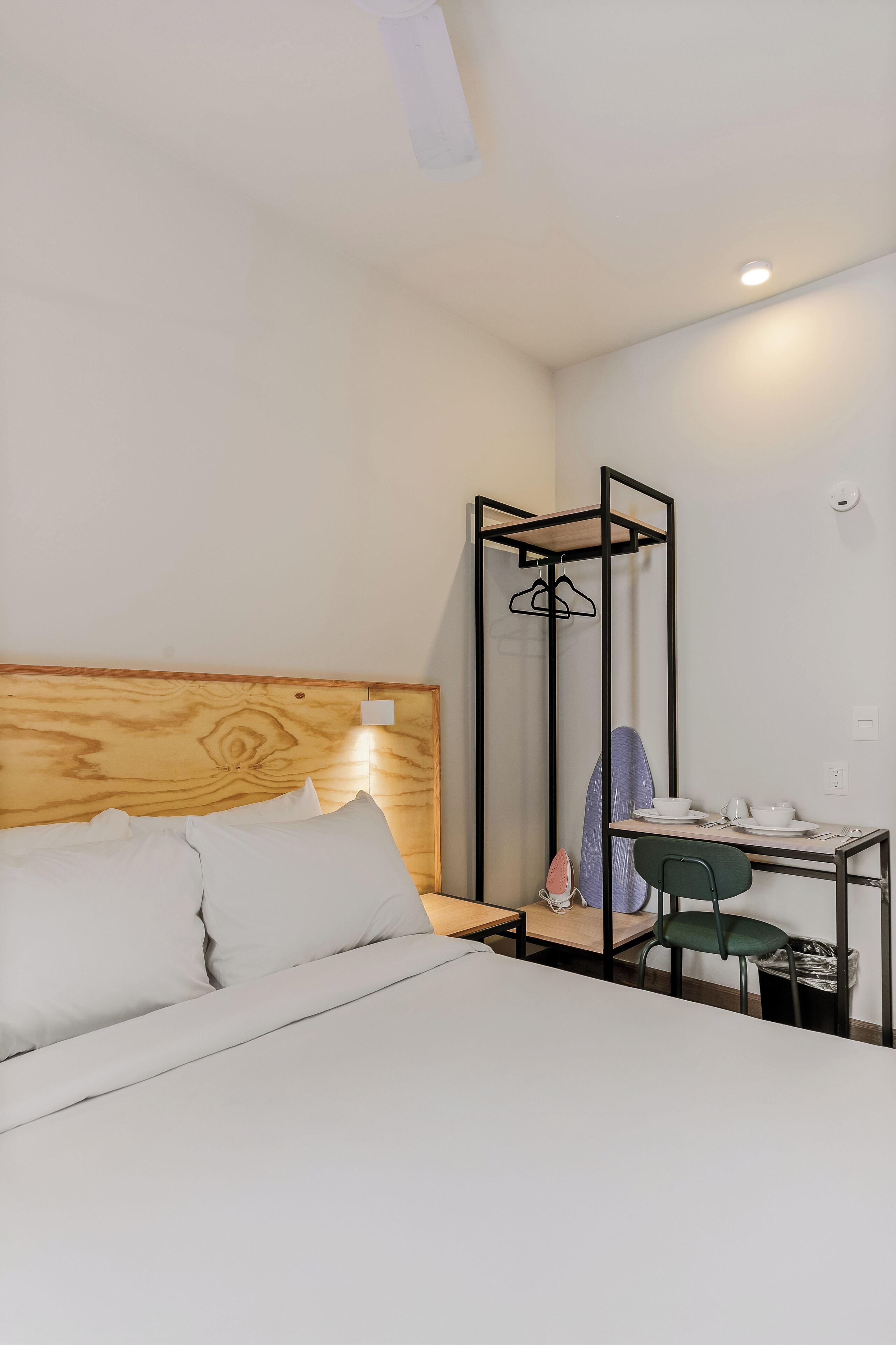 Ein Hotelzimmer mit einem hölzernen Kopfteil, einem Bett mit weißer Bettwäsche, einem Schreibtisch mit Stuhl und einem Kleiderhaken.