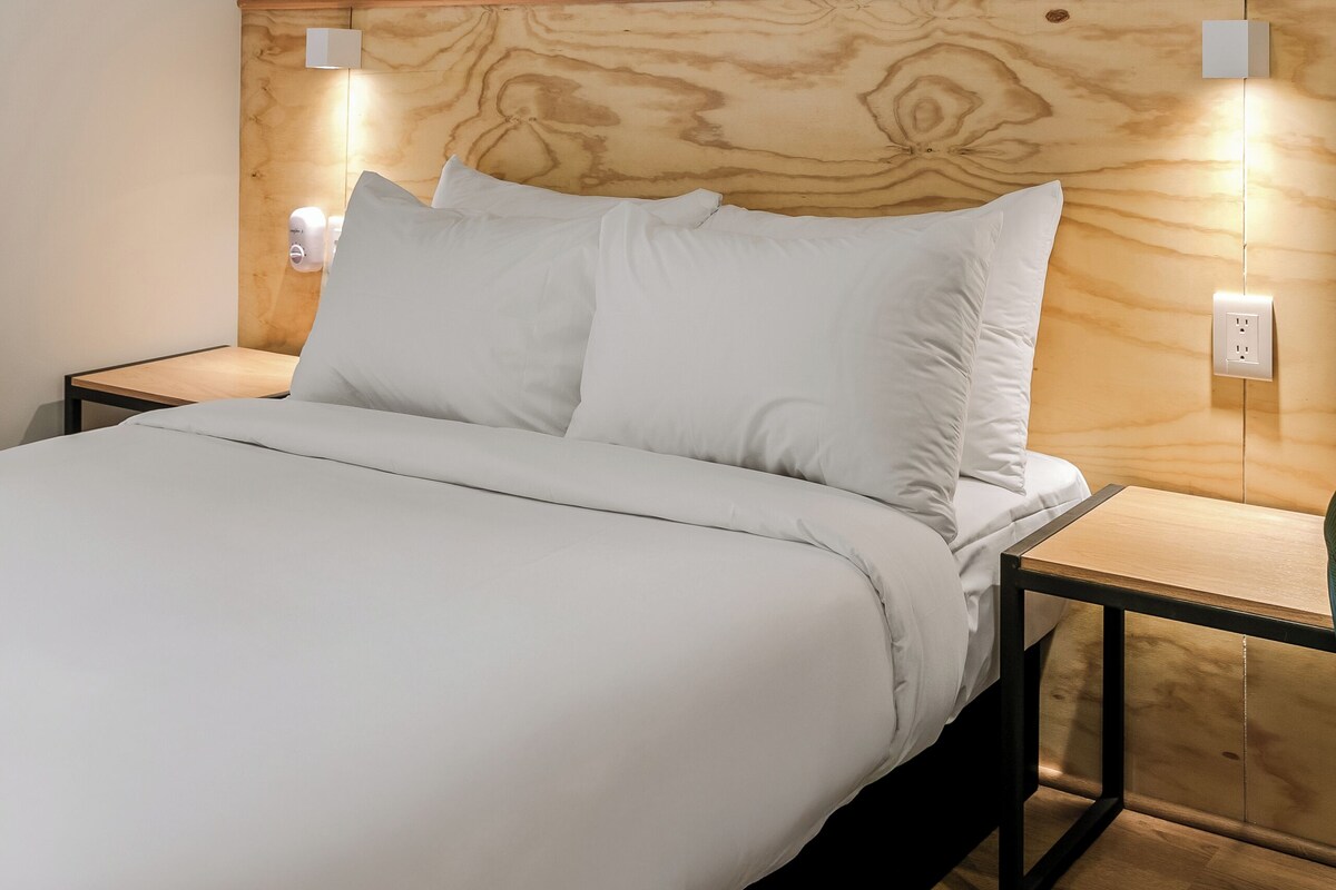 Ein ordentlich bezogenes Bett mit weißen Leinen und einem hölzernen Kopfteil. Es gibt zwei Nachttische mit an der Wand befestigten Lampen.
