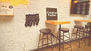 Interno di un caffè con parete in mattoni bianchi, lampadine a sospensione, insegna al neon e cartello con la scritta 'Creative Fucker'.