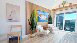 A modern living room with a wooden floor, a large window with a view of buildings, a sailboat painting, and a potted plant. - Designer Home • 1 Block to Boardwalk & Ocean • Stunning City & Ocean Views (Atlantic City)