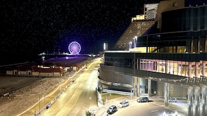 A nighttime view of a coastal area with a large Ferris wheel, a modern building with illuminated windows, and a parking lot with several cars. - Designer Home • 1 Block to Boardwalk & Ocean • Stunning City & Ocean Views (Atlantic City)