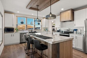 Una cocina moderna con gabinetes blancos, una isla grande con taburetes de bar, iluminación colgante y vistas al mar a través de las ventanas.