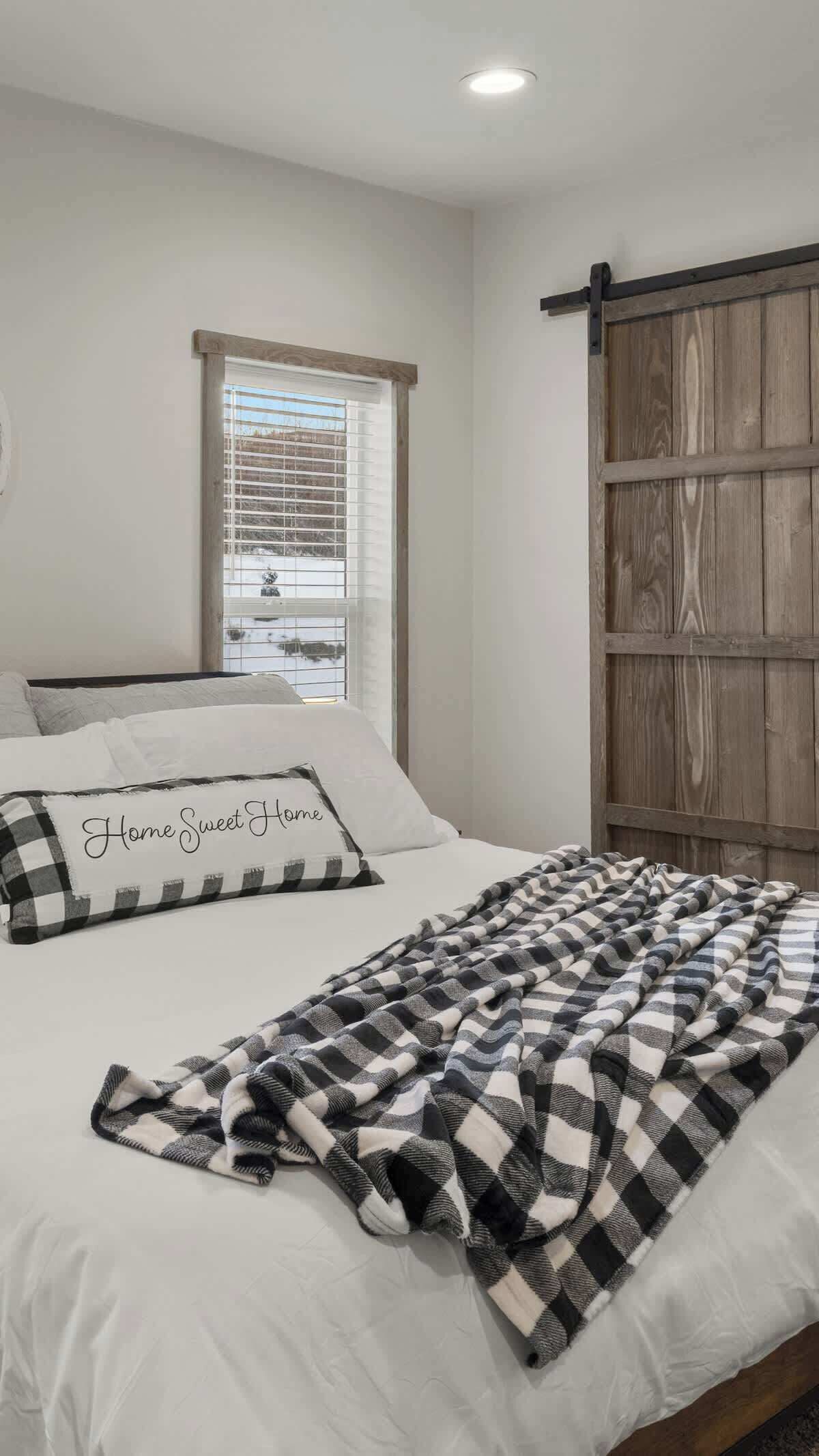 A bedroom with a bed, a window with blinds, a wooden door, and a blanket on the bed.