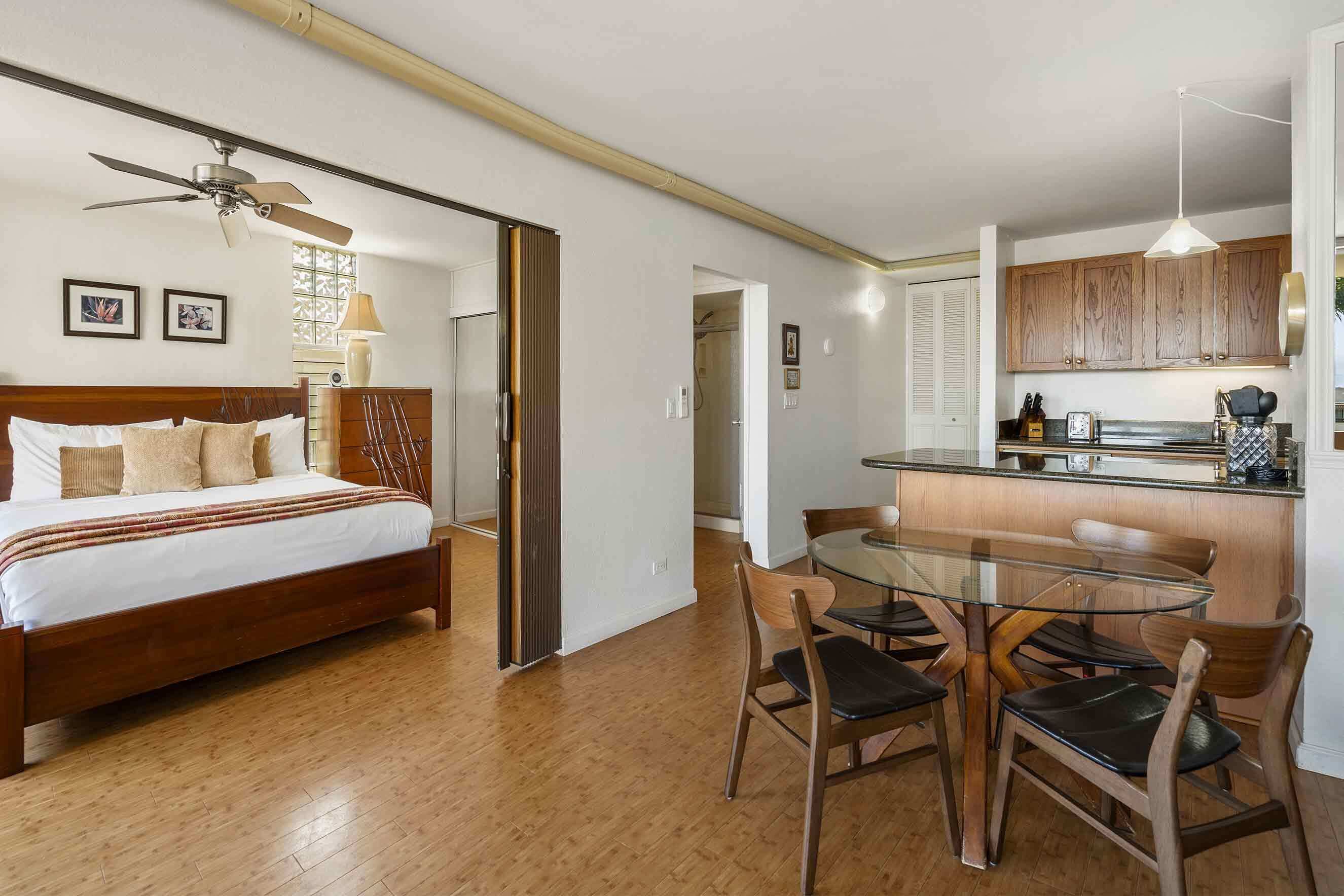 A bedroom with a wooden bed, a glass dining table with chairs, and a kitchen area with wooden cabinets and a hanging light.