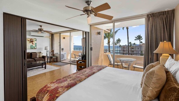 Hotelkamer met een bed, een plafondventilator, een balkon met een tafel en stoelen, en uitzicht op palmbomen en gebouwen.