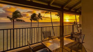Ein Balkon mit Glastisch und Stühlen, der Blick geht auf einen Sonnenuntergang über dem Meer mit Palmen.