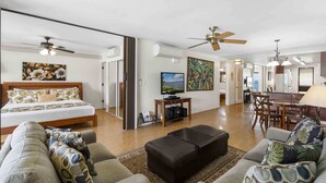A spacious living area with a sofa, ottoman, and a bed with a wooden headboard. There is a television on a stand and a ceiling fan. - Lauloa 410 - Top Floor, Oceanfront Lanai, Molokini & Halaeakala Views, Split A/C (Wailuku)