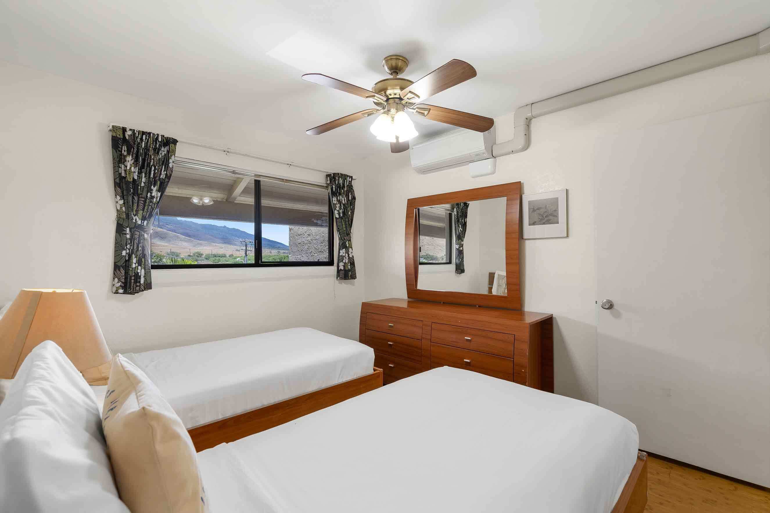 Ein Schlafzimmer mit zwei Einzelbetten, einem hölzernen Schminktisch, einem Deckenventilator, einem Fenster mit Vorhängen und einem Spiegel.