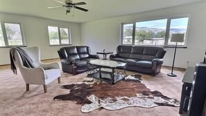 A living room with a ceiling fan, two leather sofas, a coffee table, a white armchair, and a large rug. - Full Family home on Vineyard Estate (Keremeos)