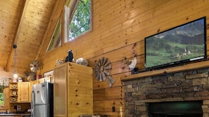 A cozy cabin interior with a stone fireplace, wooden walls, a kitchen area, and a television mounted above the fireplace.
