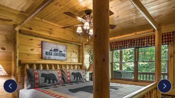 A log cabin bedroom with a bed, a sign reading "Bear Crossing", and a window with a view of trees.