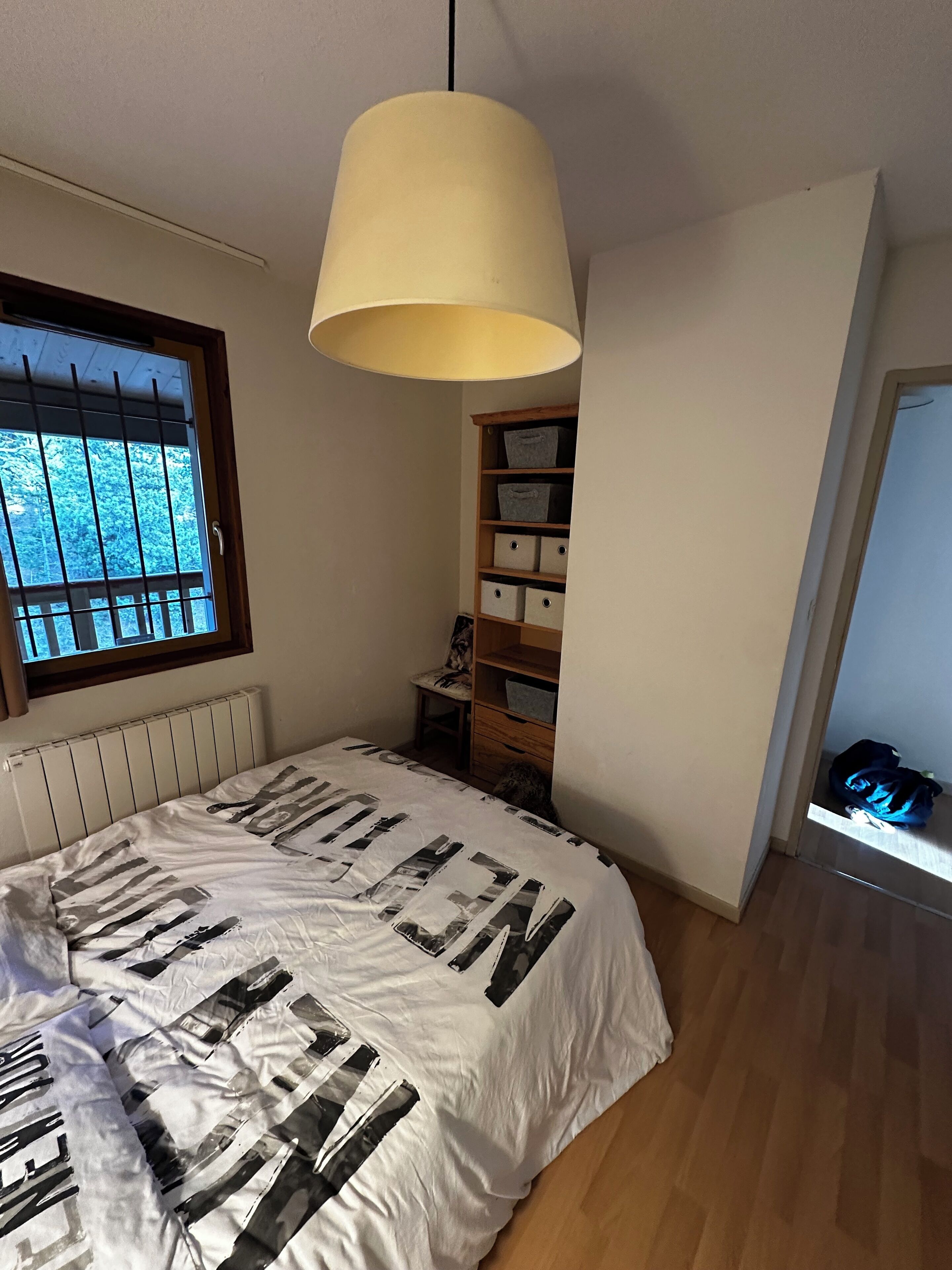 A bedroom with a bed, a window with bars, a radiator, a wooden shelf with storage boxes, and a hanging lamp.