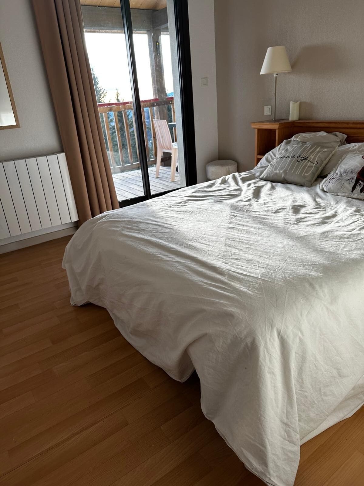 A bedroom with a large bed, a wooden floor, a sliding glass door to a balcony, and a bedside table with a lamp.
