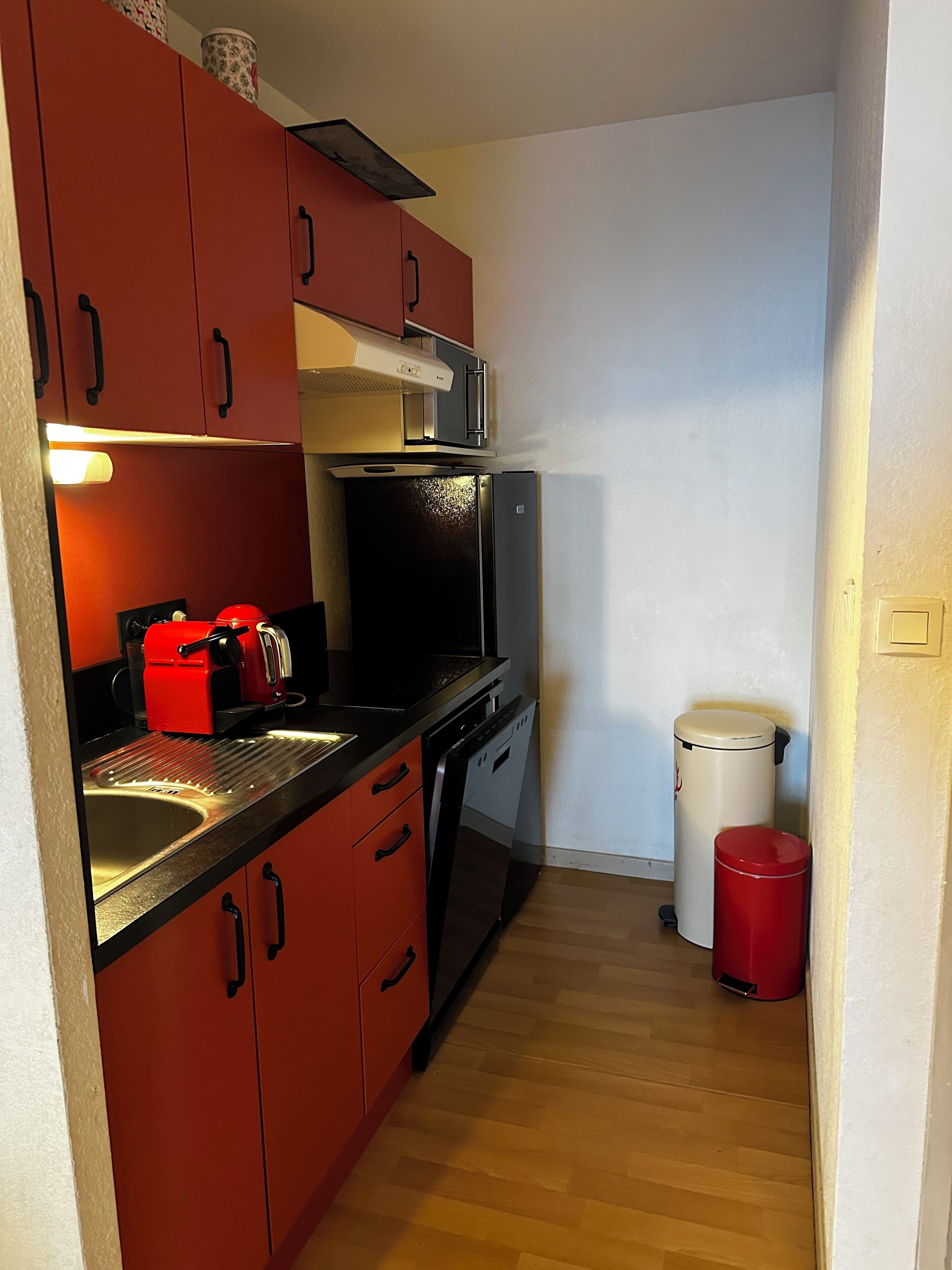 Eine kleine Küche mit roten Schränken, einem schwarzen Kühlschrank, einer Mikrowelle, einer roten Kaffeemaschine und einem weißen Mülleimer.