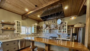 A kitchen with a wooden ceiling, hanging pots, a stainless steel refrigerator, and a bar area with stools.
