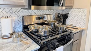 Una cocina con estufa de acero inoxidable, cuatro ollas sobre los fuegos, un microondas encima de la estufa y un salpicadero de azulejos.