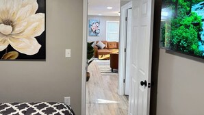 Un pasillo que conduce a una sala de estar con un cuadro grande de flores, un televisor mostrando un paisaje de bosque y una puerta a otra habitación.