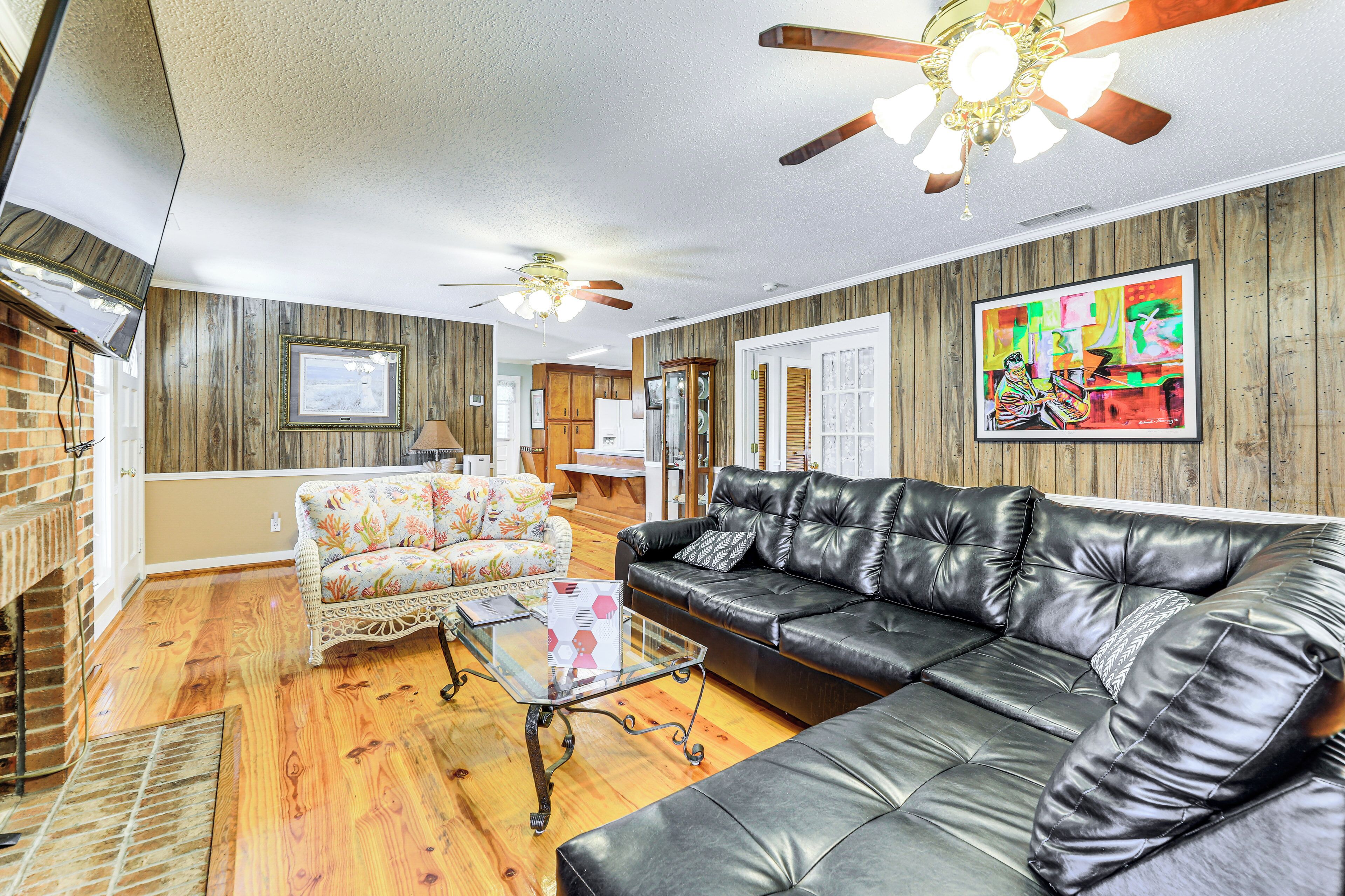 A living room with a black leather sofa, a floral armchair, and a glass coffee table. There is a ceiling fan and a painting on the wall.