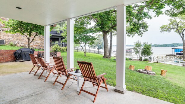 A covered patio with wooden chairs and a table, overlooking a grassy area and a body of water.