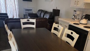 Een eetgedeelte met een donker houten tafel en witte stoelen, een zwarte leren bank, een koffiezetapparaat en een aan de muur bevestigde televisie.