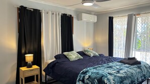 A bedroom with a bed, a ceiling fan, an air conditioning unit, a bedside lamp, and a window with curtains.