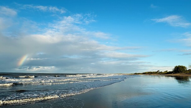 A beach with waves, a rainbow, and a clear sky.
